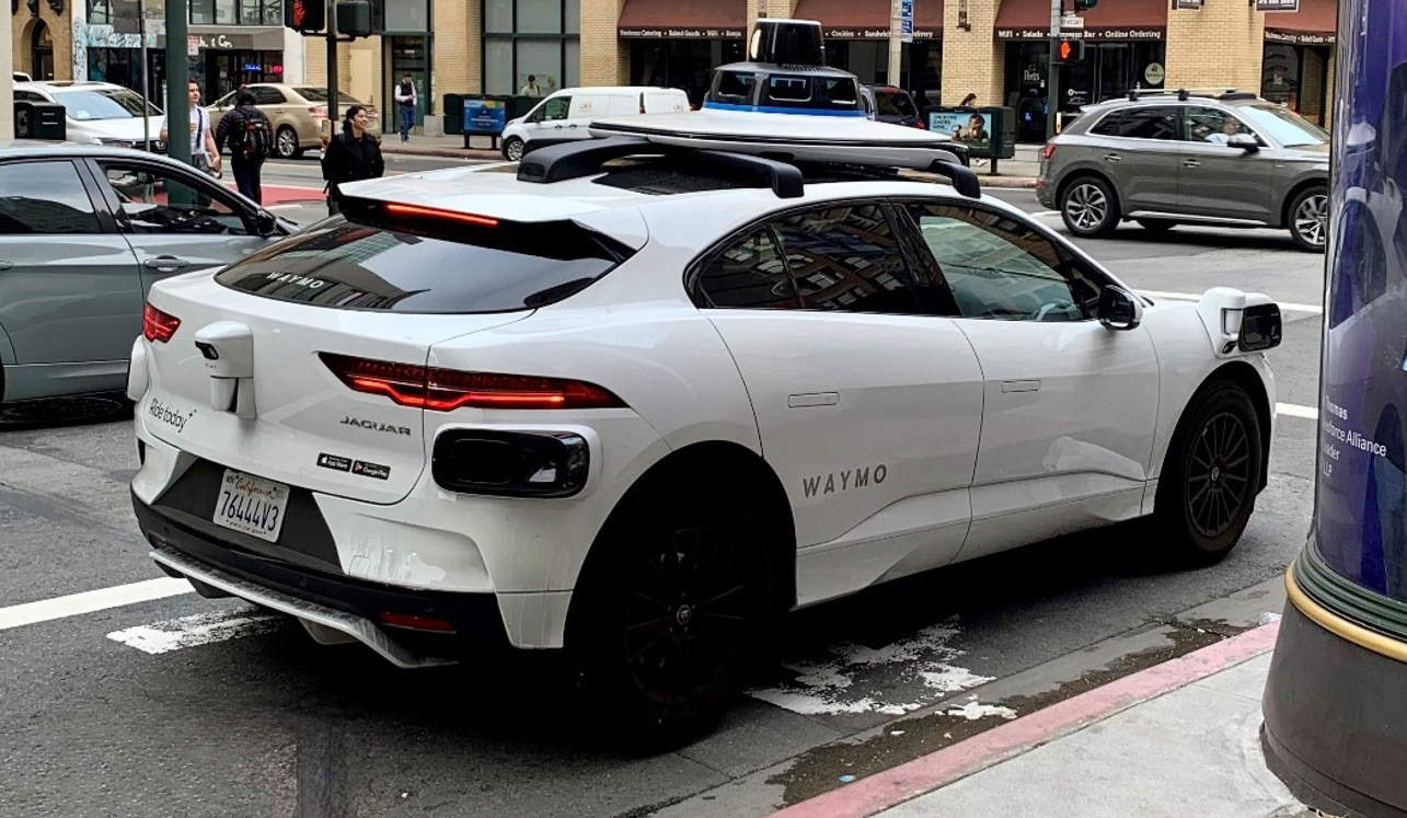 A Jaguar autonomous Waymo car waits at a cross walk in a city
