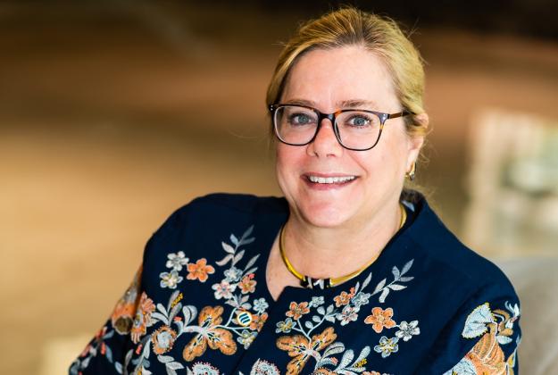 Nancy has blonde hair pulled into a low bun, wears black-rimmed glasses, and a blue floral blouse, sits in a chair and smiles at the camera