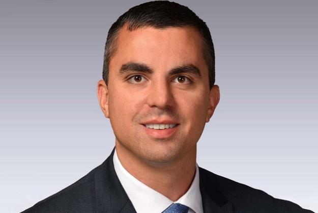 Wil wears a black suit jacket over a white dress shirt and tie and smiles at the camera