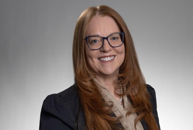 Judith wears a black jacket over a tan dress shirt, has long auburn hair and wears glasses