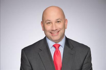 Ryan Hanna wears a black suit jacket, red tie, and smiles at the camera