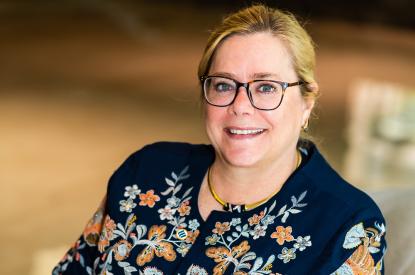 Nancy has blonde hair pulled into a low bun, wears black-rimmed glasses, and a blue floral blouse, sits in a chair and smiles at the camera