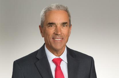 Jeff wears a black suit jacket over a white shirt and red tie and smiles at the camera