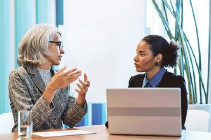 Two women are having a conversation at a desk. One of them is using a laptop.