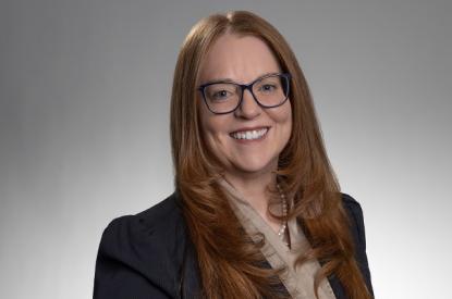 Judith wears a black jacket over a tan dress shirt, has long auburn hair and wears glasses